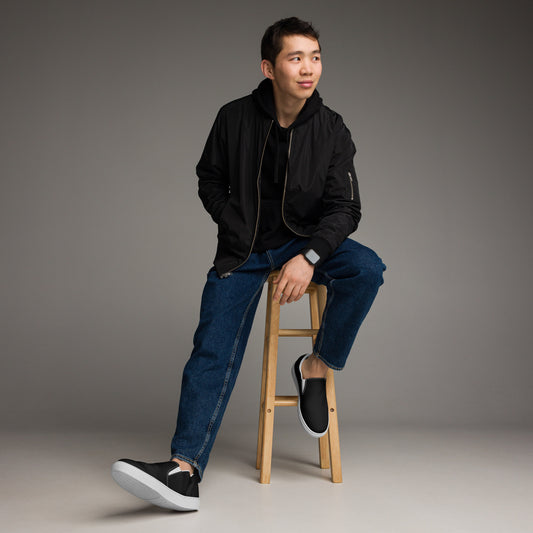 A man sitting on a stool wearing black slip-on canvas shoes with white soles.