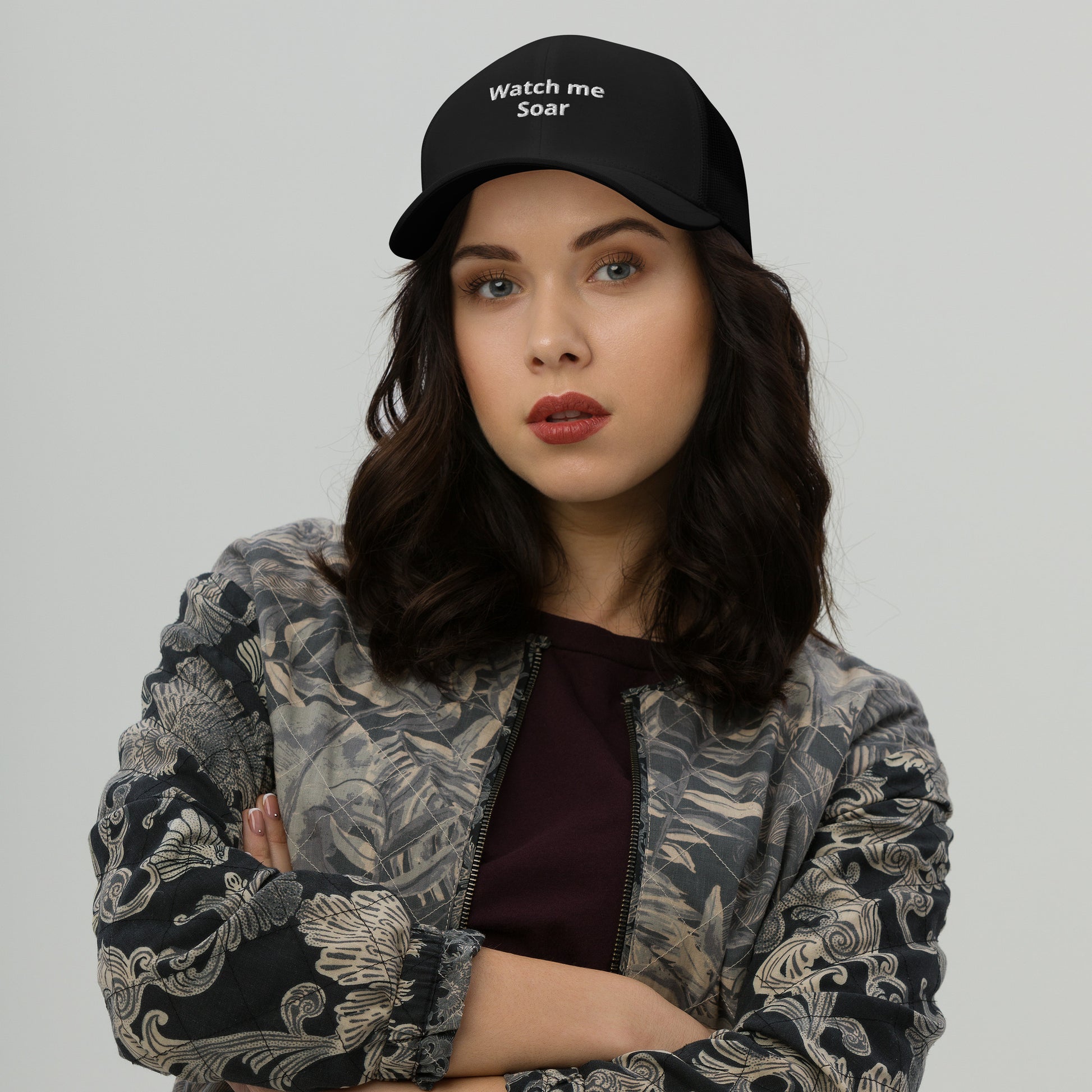 A woman wearing a black trucker cap with the text 'watch me Soar' on the front.
