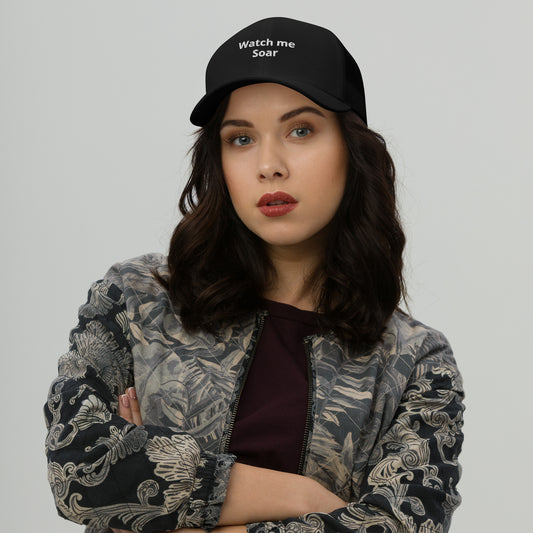 A woman wearing a black trucker cap with the text 'watch me Soar' on the front.