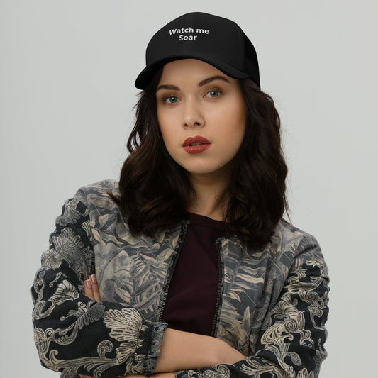A woman wearing a black trucker cap with the text 'watch me Soar' on the front.