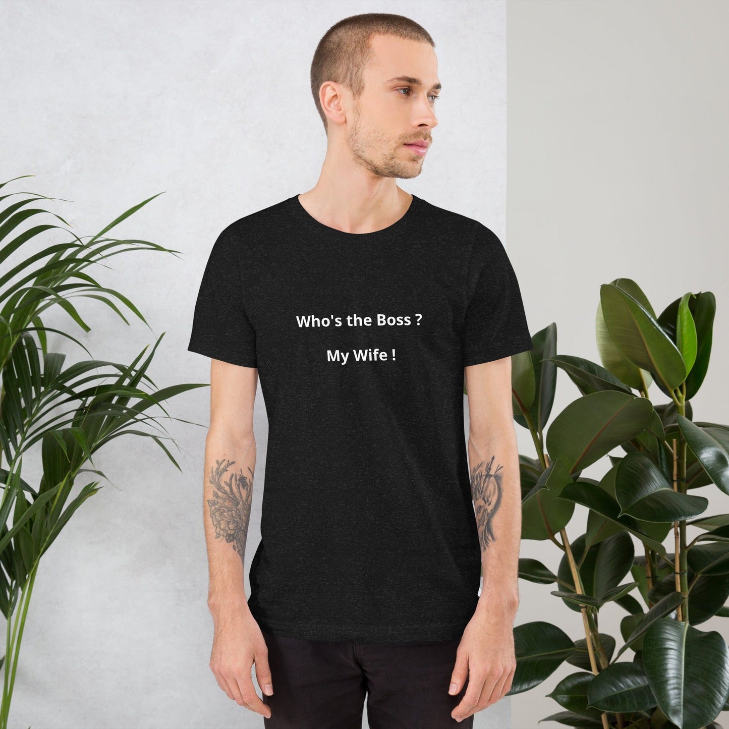 A man wearing a black t-shirt with the text 'Who's the Boss? My Wife!' standing indoors among plants.